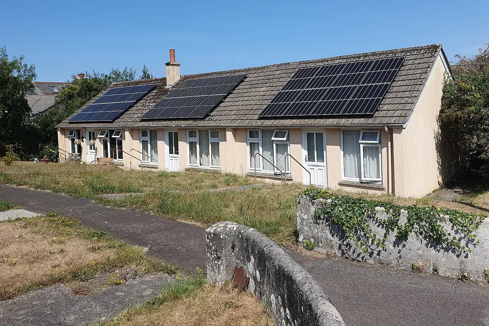 Three solar panel arrays on the tiled roof of a row of rendered bungalows, seen from the pavement on a sunny day