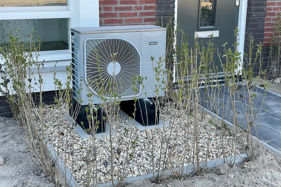Air source heat pump outdoor unit on a gravel bed beside the brick wall and front door of a house