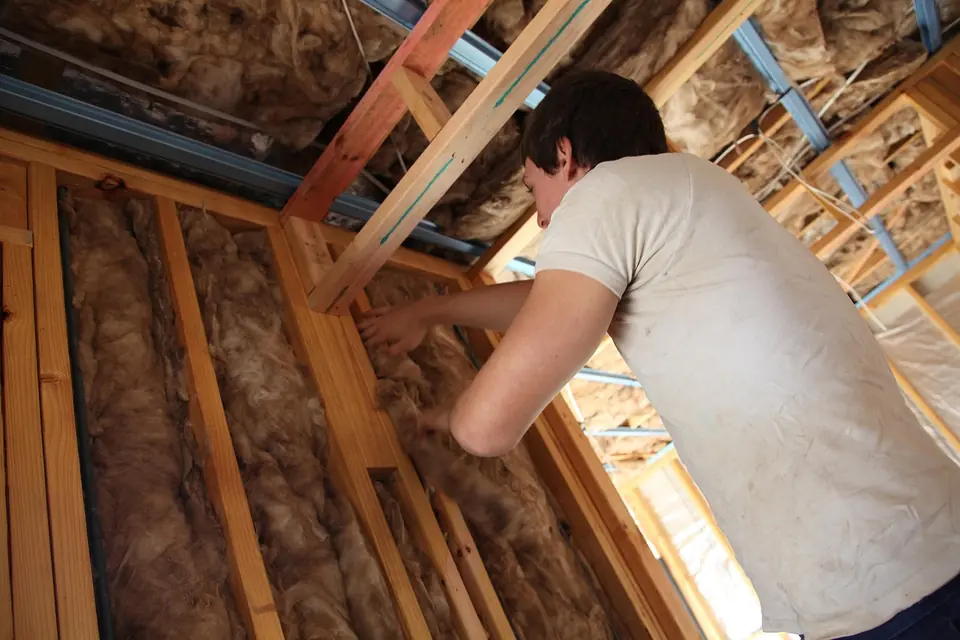 Person laying mineral wool insulation between the timber joists of a loft floor, seen from behind