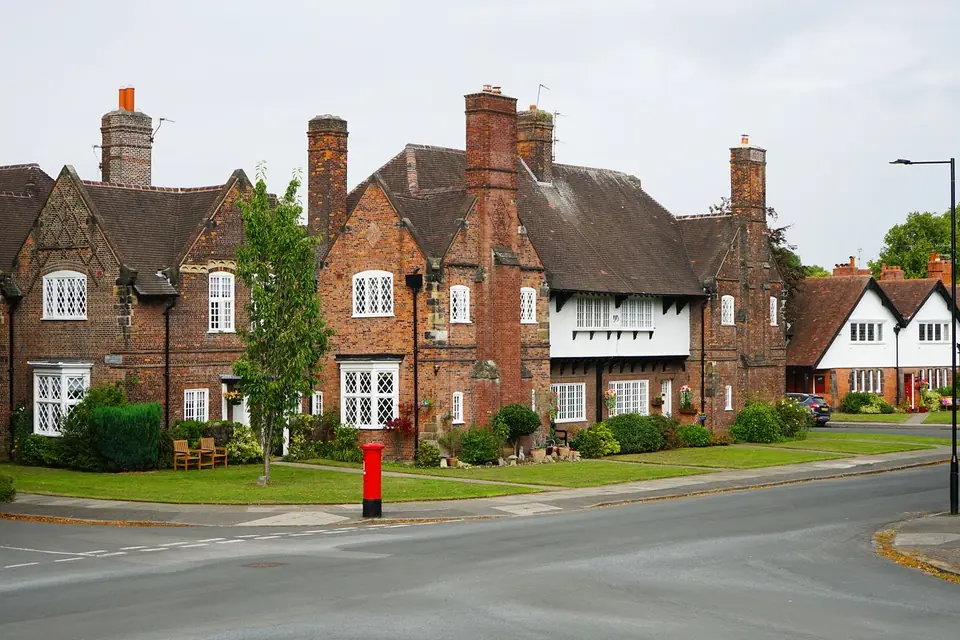 Row of red brick cottages with tall chimneys and white windows on a street corner, beside a grass verge and a red postbox