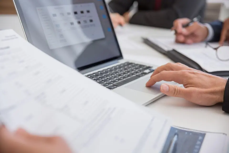 Hands reviewing printed documents beside an open laptop on a white table, with a pen and clipboard
