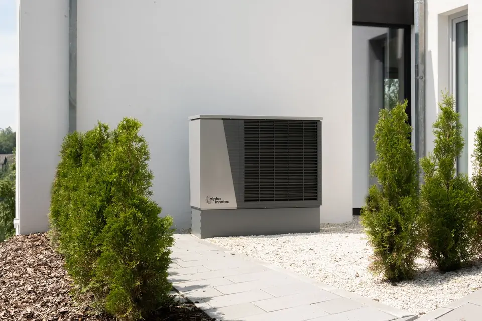 Grey outdoor air source heat pump unit standing against the white rendered wall of a modern house beside small conifers
