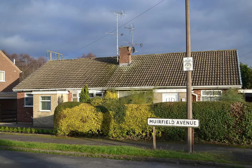 Semi-detached brick and stone bungalow with a tiled roof, chimney and clipped front hedge on a suburban street