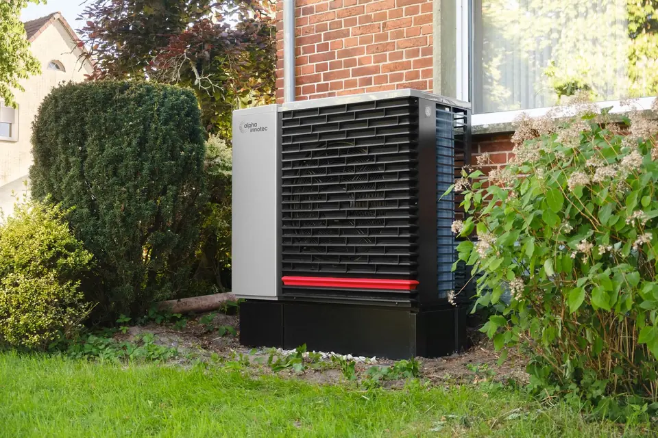 Air source heat pump with a large front fan on a plinth beside the red brick wall of a house, framed by garden shrubs
