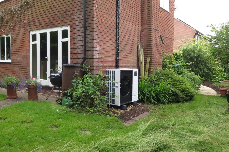 White air source heat pump beside a red brick house, with a garden hedge and neighbouring house behind