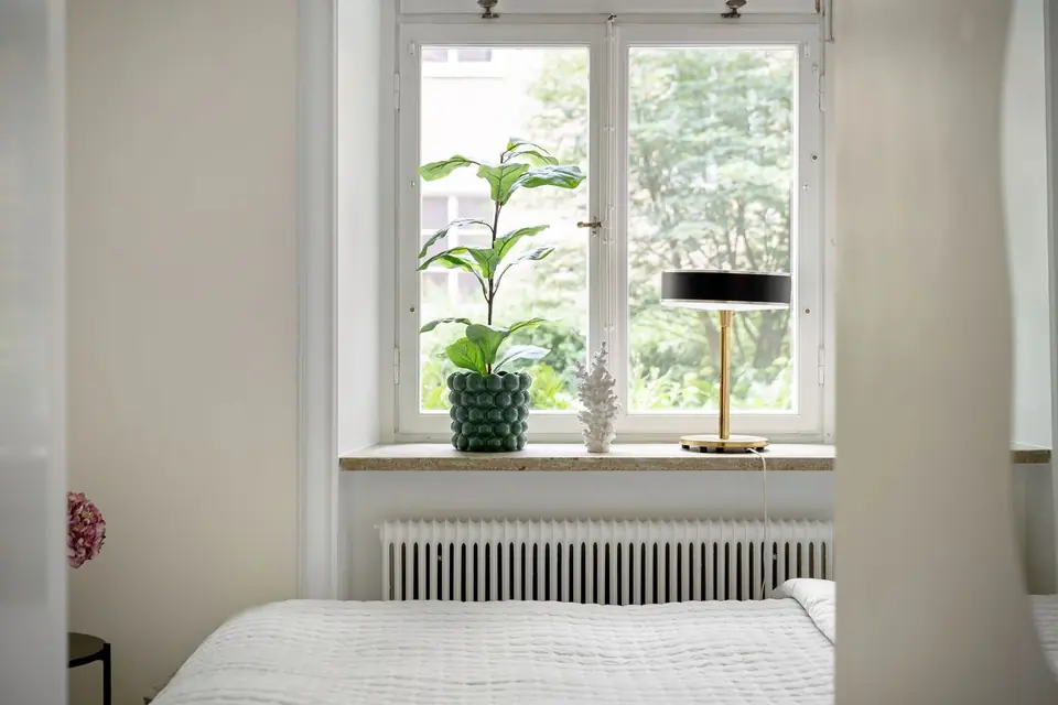 White radiator fitted under a bedroom window, with a potted plant and lamp on the sill and a bed in front