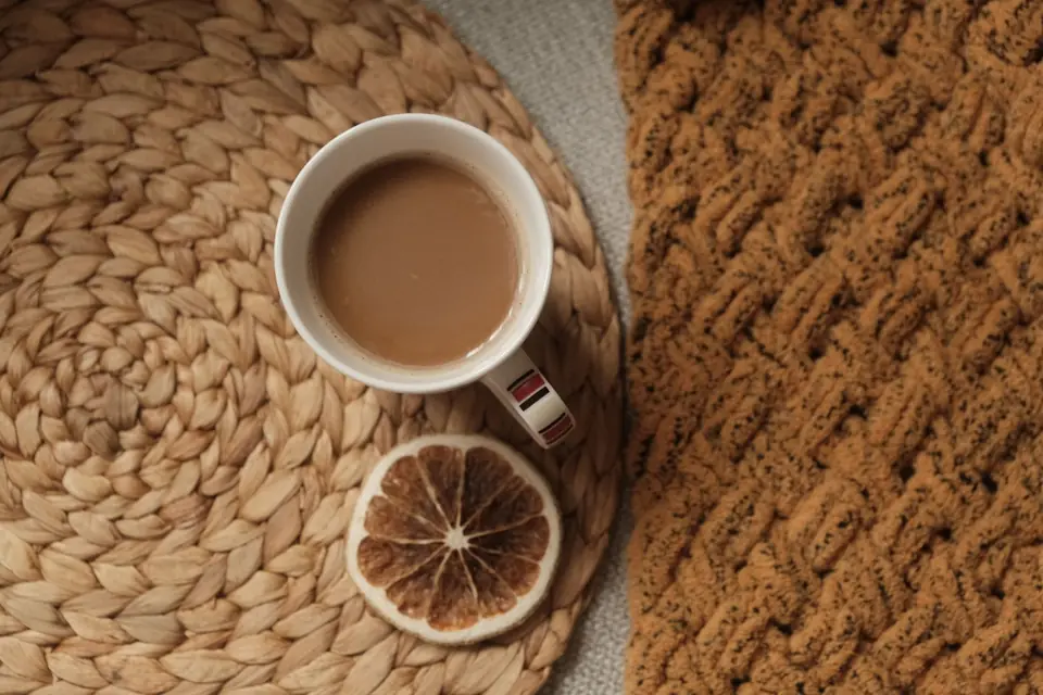 Mug of milky tea on a woven mat beside a thick knitted blanket in a warm living room